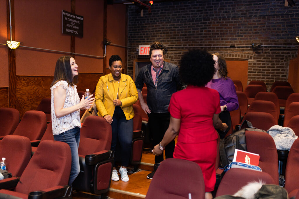 A group of people are standing in a circle in a theater with red seats. They are laughing and smiling