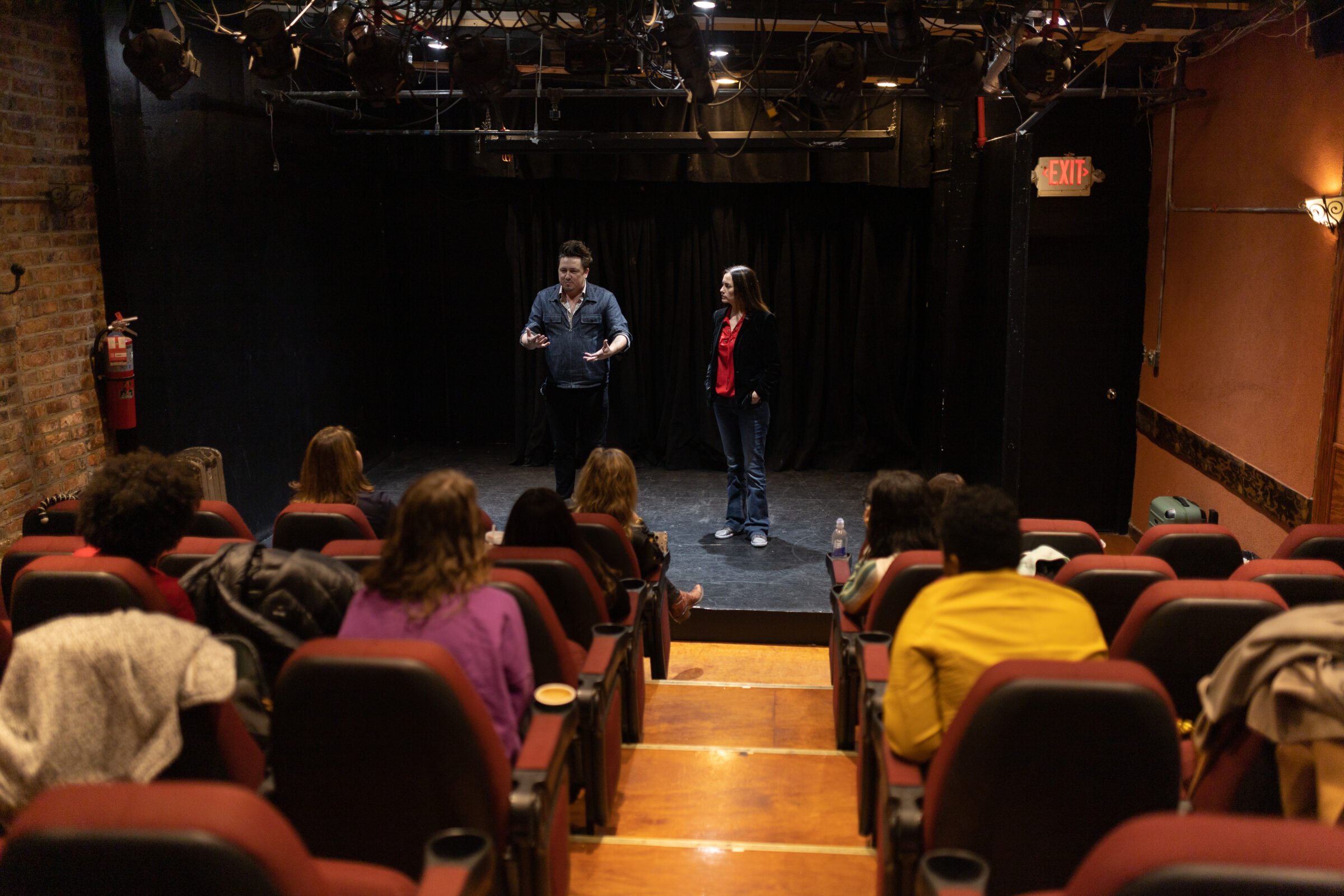 An image from the back of a small theater. People are in the audience. Two people are on stage. One of them is teaching.